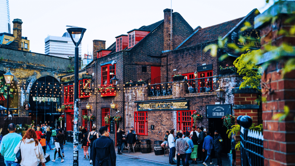 The Anchor Bankside, a historic 17th-century brick pub with iconic red window frames and string lights, crowded with people during the evening on London's South Bank.
